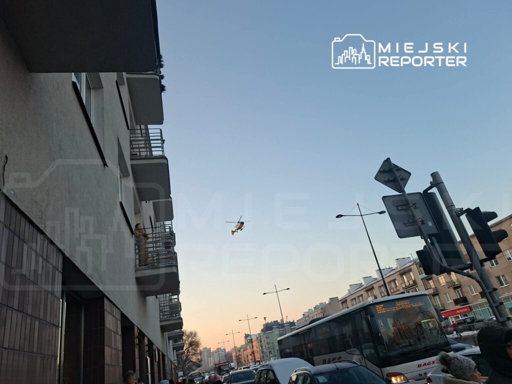 Kobieta na balkonie obserwująca helikopter przelatujący nad ulicą w zabudowanej okolicy.