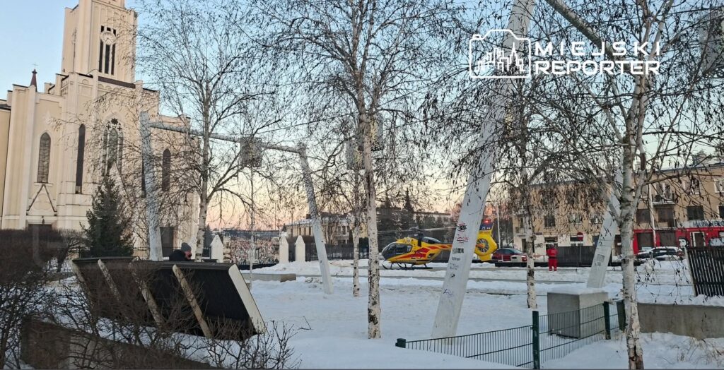 Mężczyzna w szarej czapce siedzi na ławce, obok ląduje helikopter ratunkowy na śnieżnym placu.
