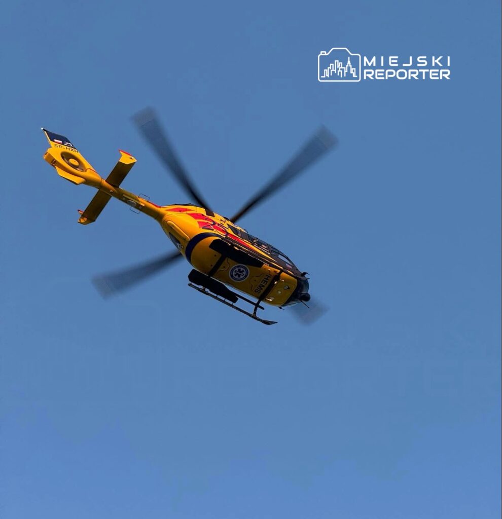 Żółto-czerwony śmigłowiec ratunkowy przelatuje nad miastem na jasnym niebie.