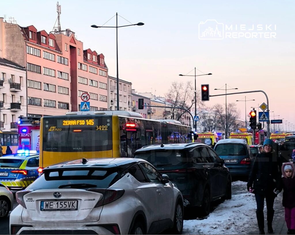 Ruchliwa ulica z autobusem miejskim, zaparkowanymi samochodami oraz pojazdami służb ratunkowych w tle.
