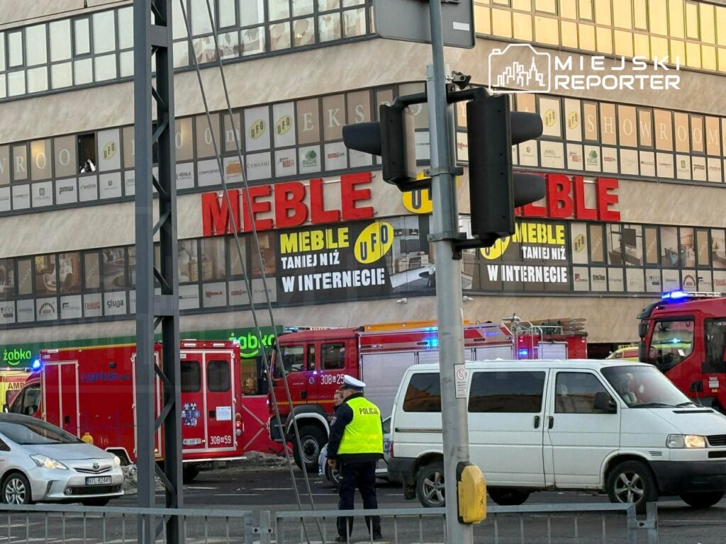 Czerwone wozy strażackie i policjant w kamizelce ostrzegawczej na parkingu przed budynkiem ze sklepem meblowym.