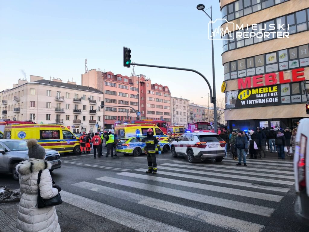 Grupa ratowników i policjantów pracuje na skrzyżowaniu, obok karetek pogotowia i samochodów służbowych.