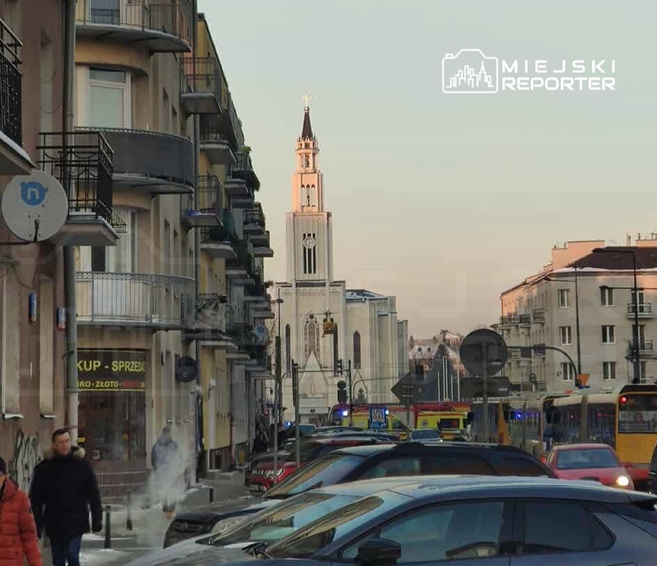 Ulica z zaparkowanymi samochodami, w tle widać kościół z wysoką wieżą i krzyżem, a po bokach budynki mieszkalne.