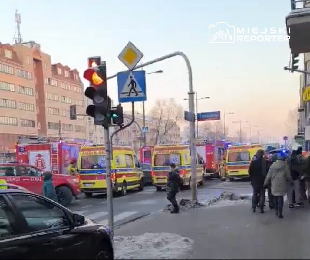 Na skrzyżowaniu ulic parkują karetki pogotowia i samochody straży pożarnej, a przechodnie obserwują sytuację.