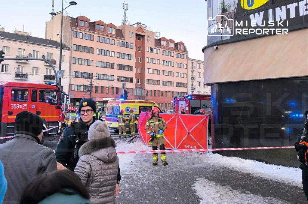 Grupa strażaków i ratowników medycznych pracuje przy wypadku w miejskim otoczeniu, zabezpieczonym taśmą.