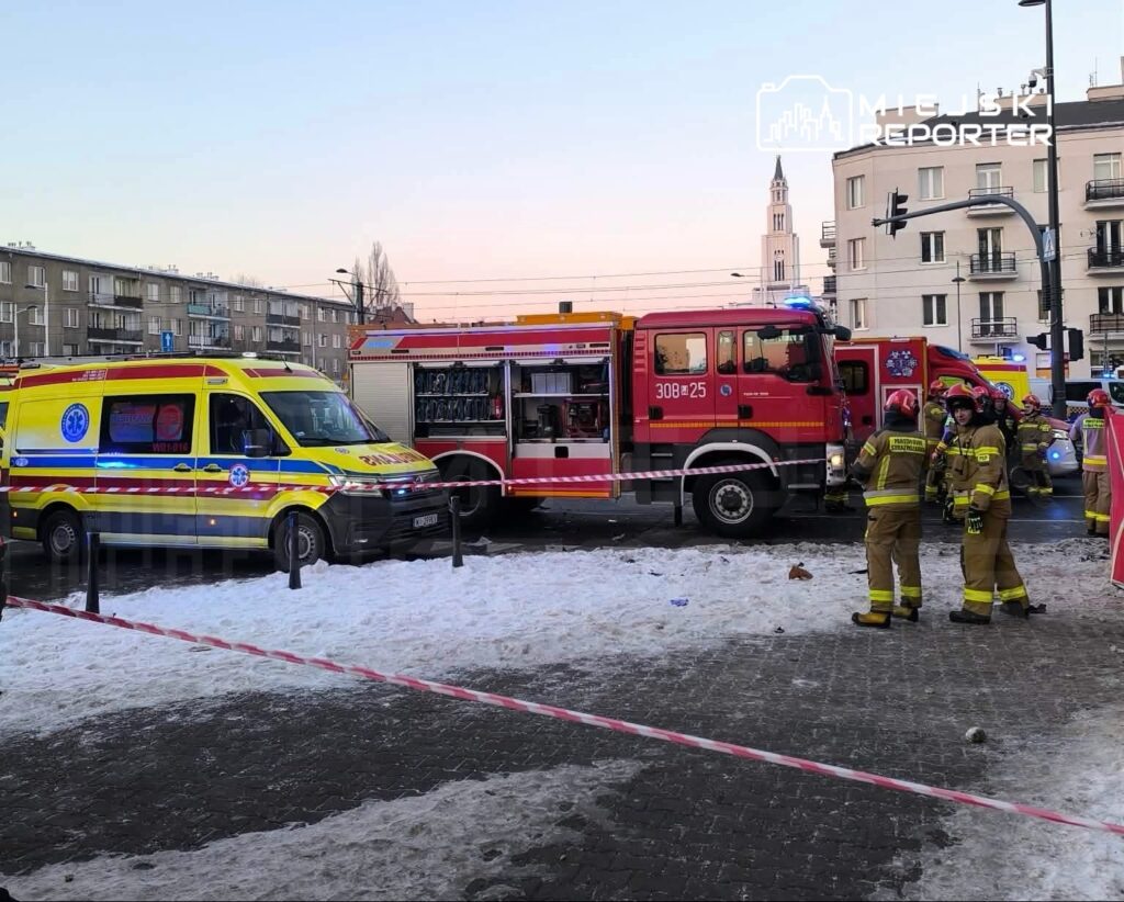 Na ulicy stoją karetka pogotowia i wóz strażacki, a strażacy w mundurach rozmawiają obok. W tle widać budynki.