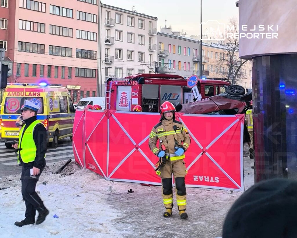 Ratownicy w mundurach pracują przy wypadku drogowym, obok przewróconego samochodu i pojazdów służb ratunkowych.