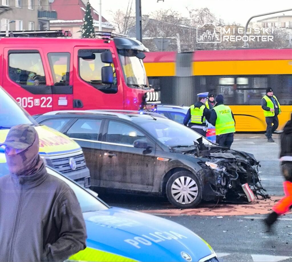 Na skrzyżowaniu zatrzymane pojazdy, w tym uszkodzony samochód, obok interweniujący policjanci i straż pożarna.