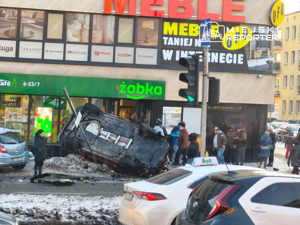 Samochód przewrócony na dach leży na chodniku przed sklepem Żabka, a obok grupka ludzi obserwuje sytuację.