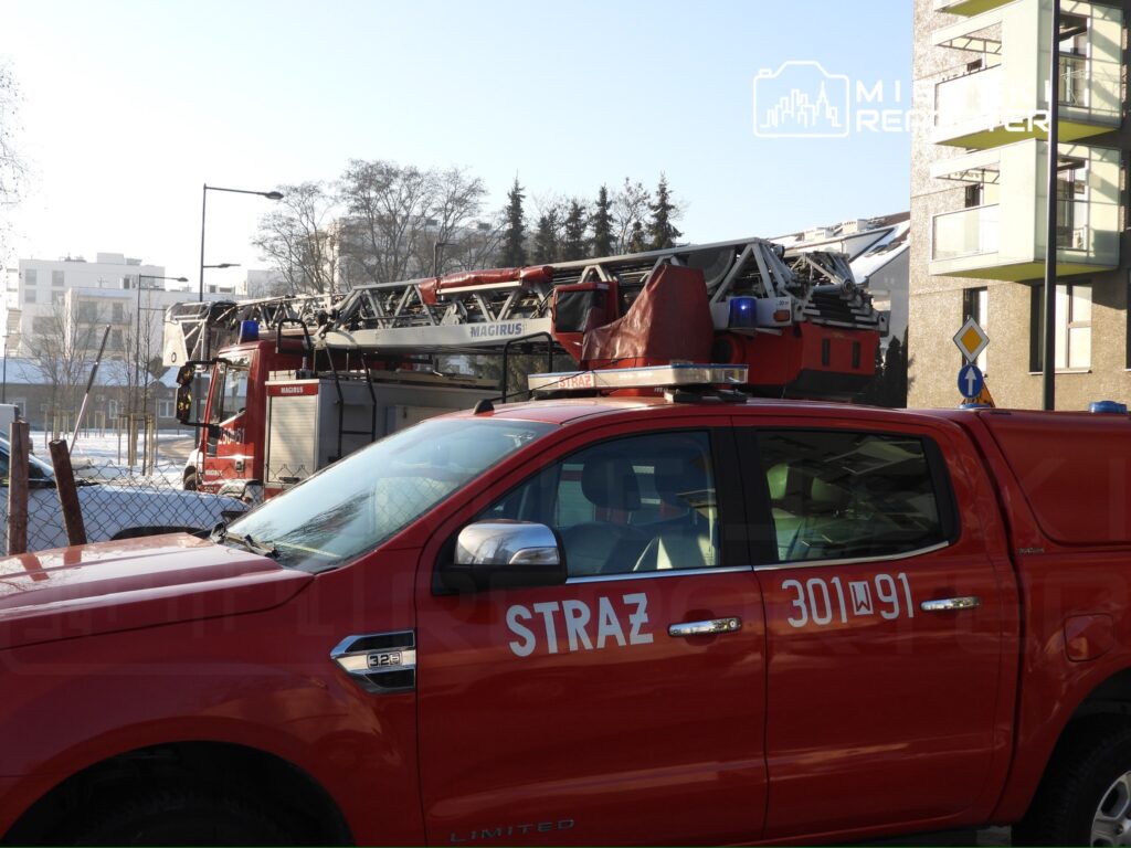 Czerwony wóz strażacki z podnośnikiem na ulicy, obok zaparkowany samochód straży pożarnej. W tle budynki mieszkalne.