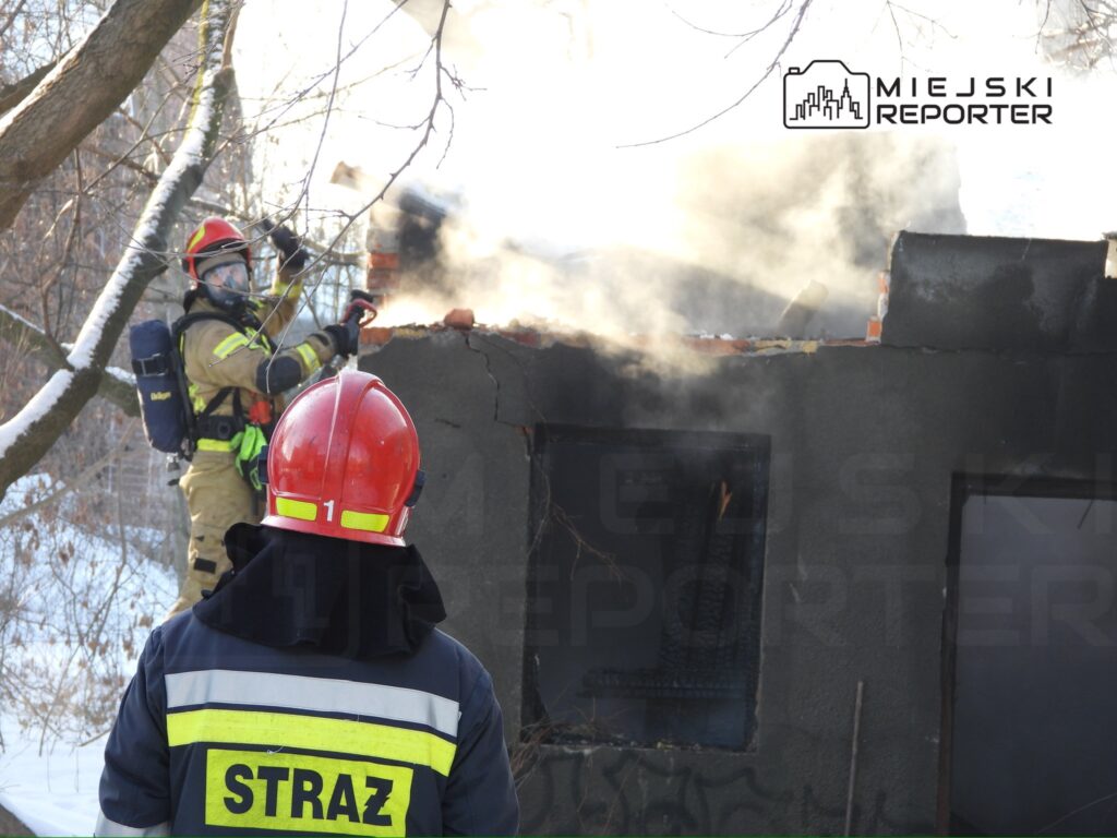 Strażak w hełmie i mundurze gasi pożar, usuwając fragmenty dachu budynku, z którego wydobywa się dym.