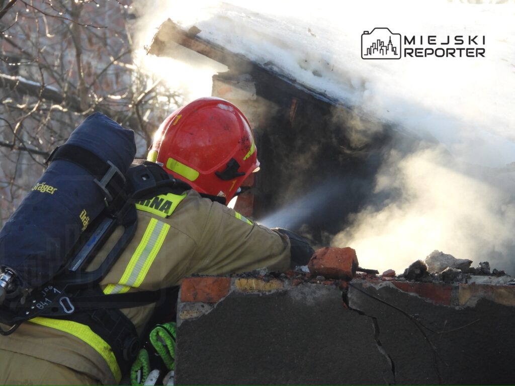 Strażak w hełmie i kombinezonie gaśniczym polewa wodą dymiący fragment dachu budynku.