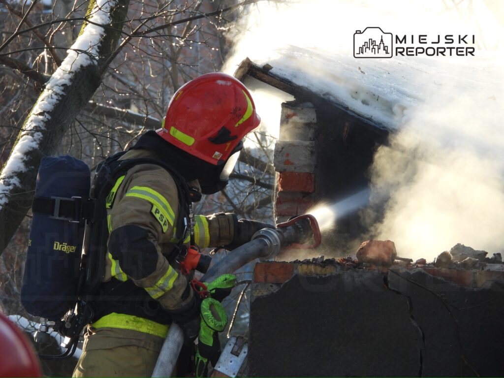 Strażak w hełmie i mundurze używa węża gaśniczego do gaszenia ognia w zniszczonym budynku. W tle widać dym.