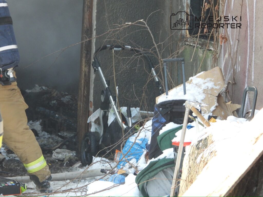 Strażak w mundurze przeszukuje zgliszcza przy zniszczonym budynku, w tle wózek dziecięcy i resztki odpadów.