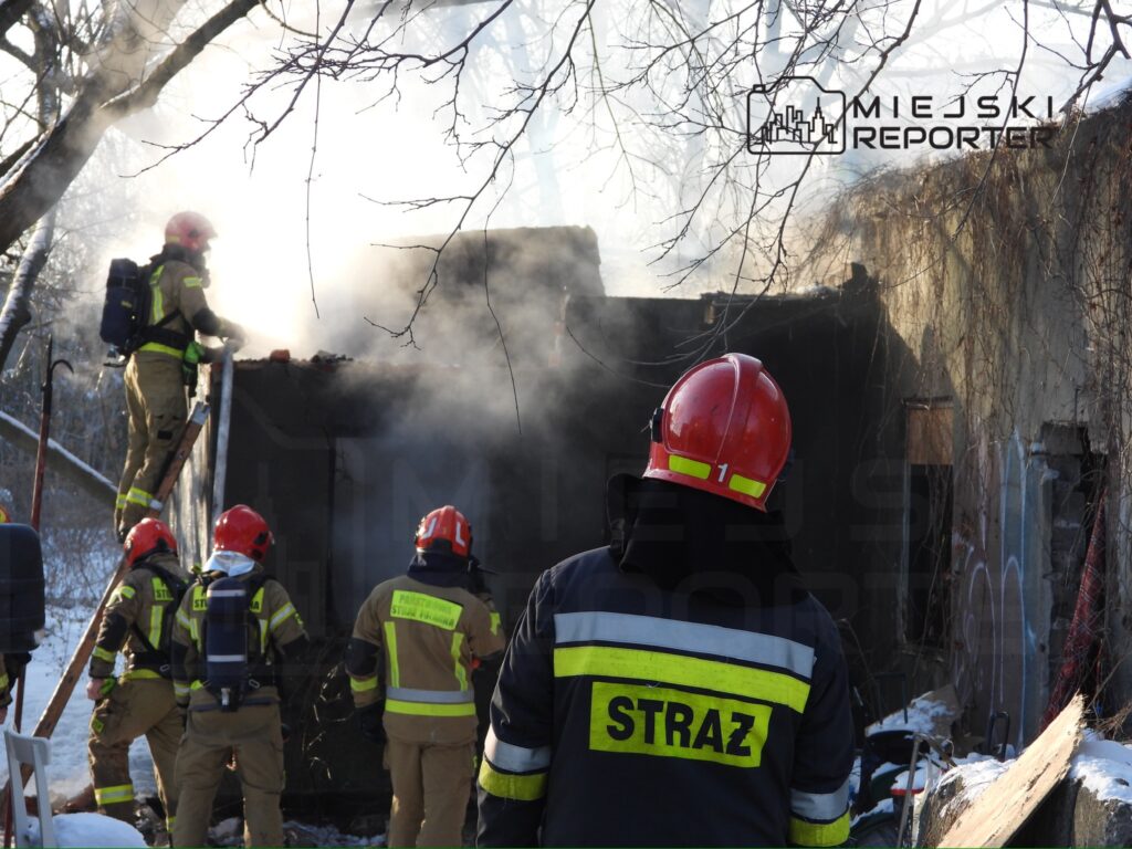 Strażacy w hełmach gaszą pożar budynku, z którego wydobywa się dym, w otoczeniu zimowej scenerii.