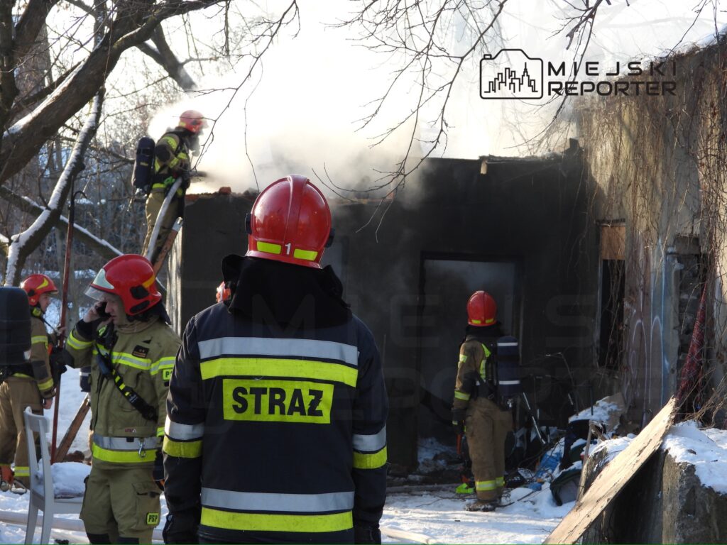 Strażacy w hełmach przeszukują zgliszcza budynku, z którego wydobywa się dym, w otoczeniu pokrytym śniegiem.