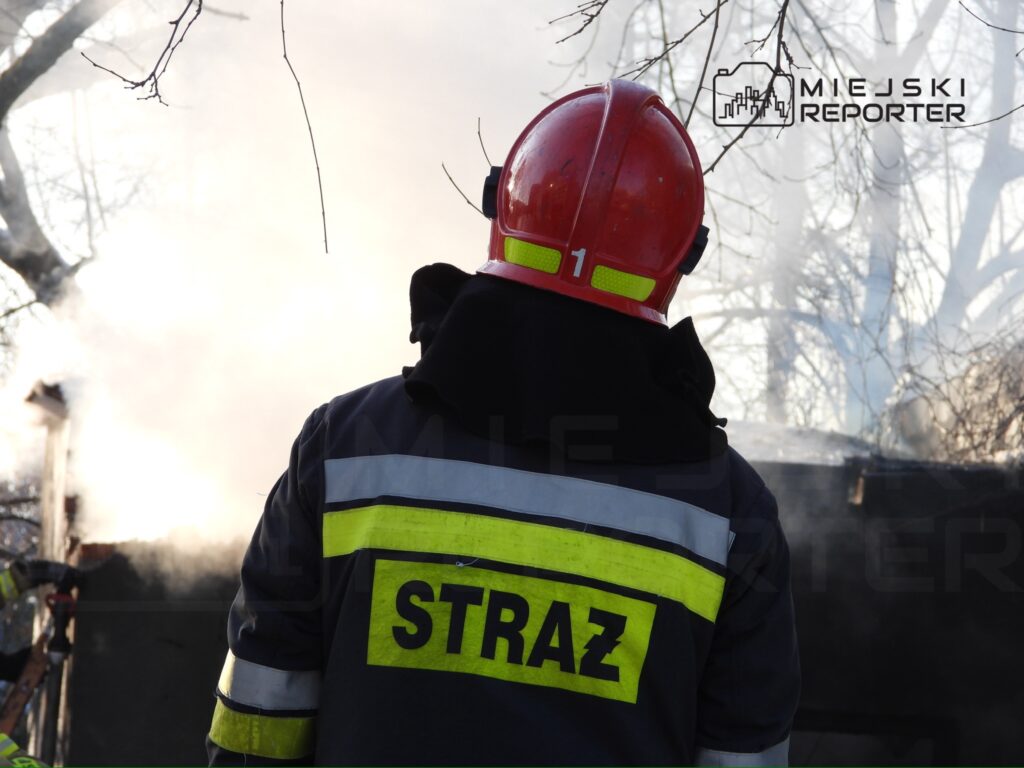 Strażak w hełmie obserwujący pożar, w tle unoszący się dym z płonącego budynku.