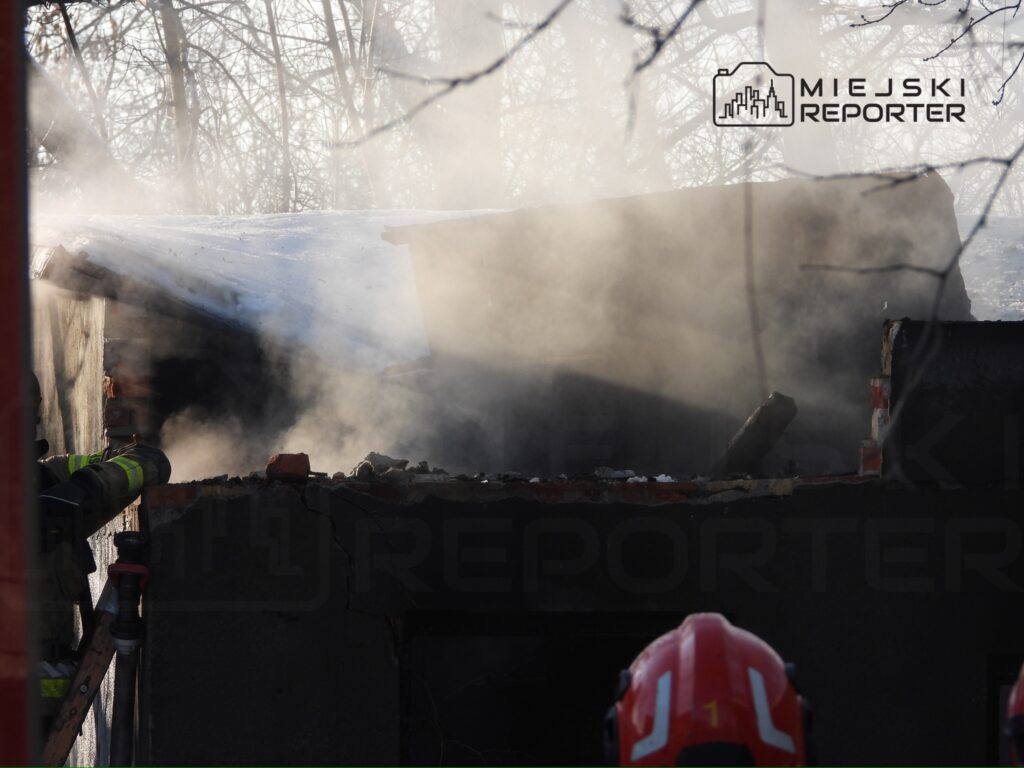Strażak w hełmie kieruje strumień wody na palący się dach, z którego unosi się gęsty dym.