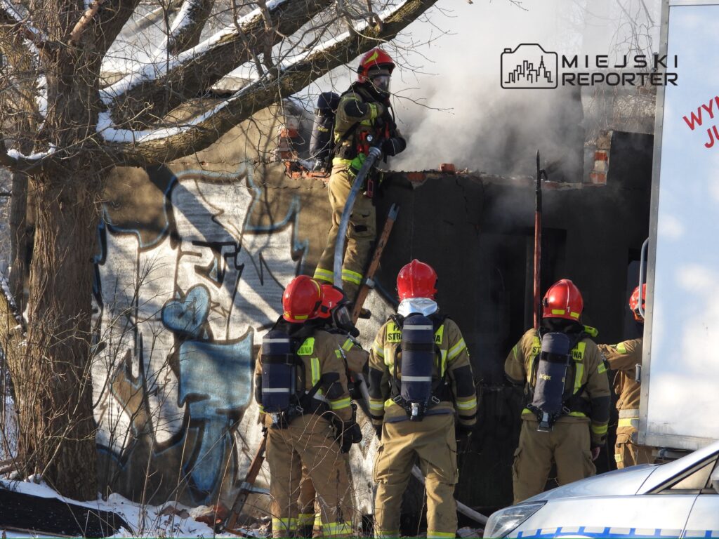 Grupa strażaków w hełmach i specjalnym ubraniu gasi pożar w zniszczonym budynku, z którego wydobywa się dym.