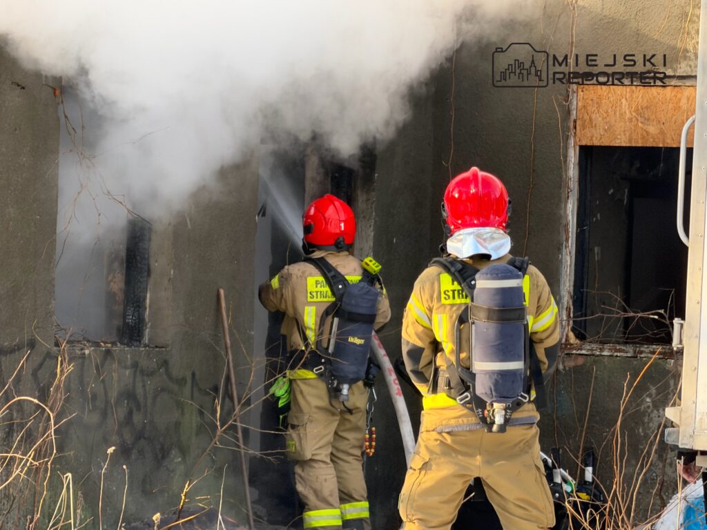 Dwaj strażacy w hełmach gaszą ogień w zadymionym budynku, używając węża strażackiego.