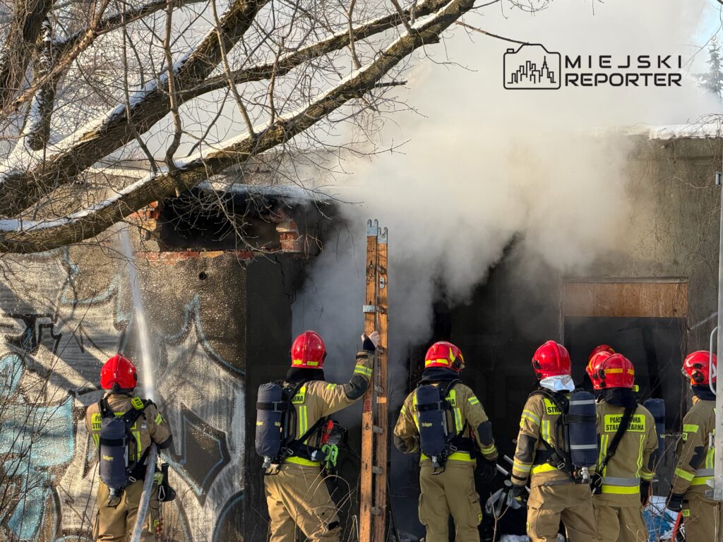 Grupa strażaków w hełmach i specjalnych strojach ugasza pożar w budynku, z którego wydobywa się dym.