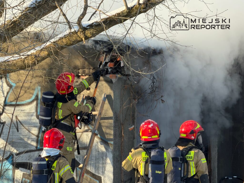 Czterech strażaków w hełmach i mundurach gaśniczych walczy z ogniem, używając narzędzi w zadymionym otoczeniu.
