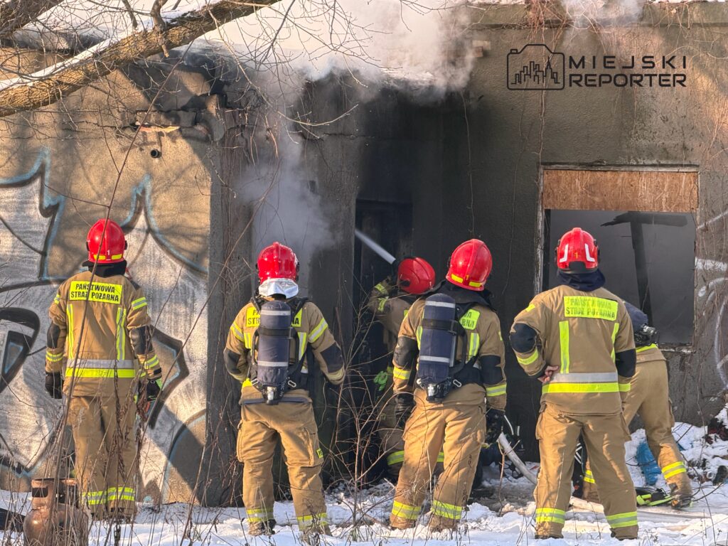 Grupa strażaków w hełmach z czerwonymi czapkami gasi pożar w opuszczonym budynku, z którego wydobywa się dym.