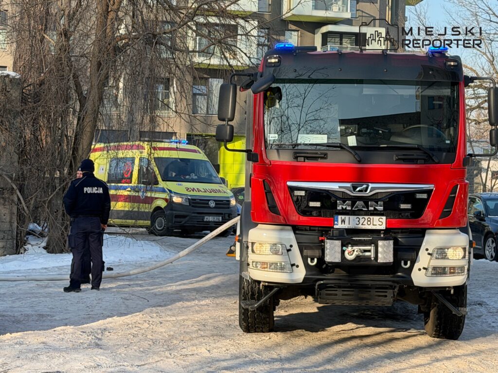 Policjant rozmawia z ratownikami medycznymi obok czerwonego wozu strażackiego na zaśnieżonej ulicy.
