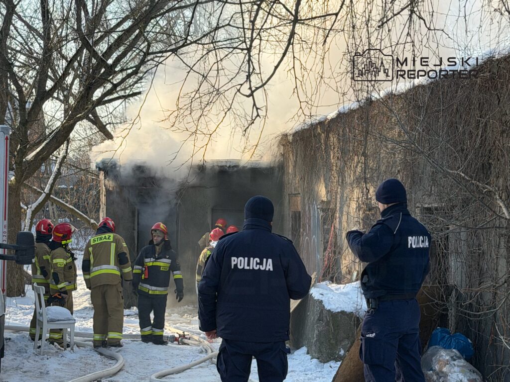 Grupa strażaków w mundurach gaśniczych otacza budynek, z którego wydobywa się dym, podczas akcji ratunkowej w zimowym otoczeniu.
