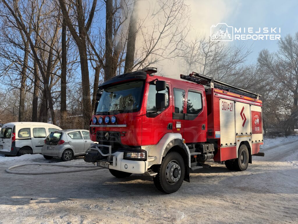 Czerwony wóz strażacki z włączonymi sygnalizatorami stoi na zaśnieżonej ulicy, obok zaparkowane samochody.