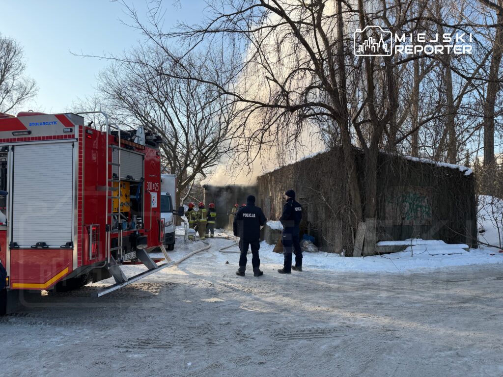 Strażacy gaszą pożar w drewnianym budynku, a obok nich stoją policjanci w zimowym otoczeniu.
