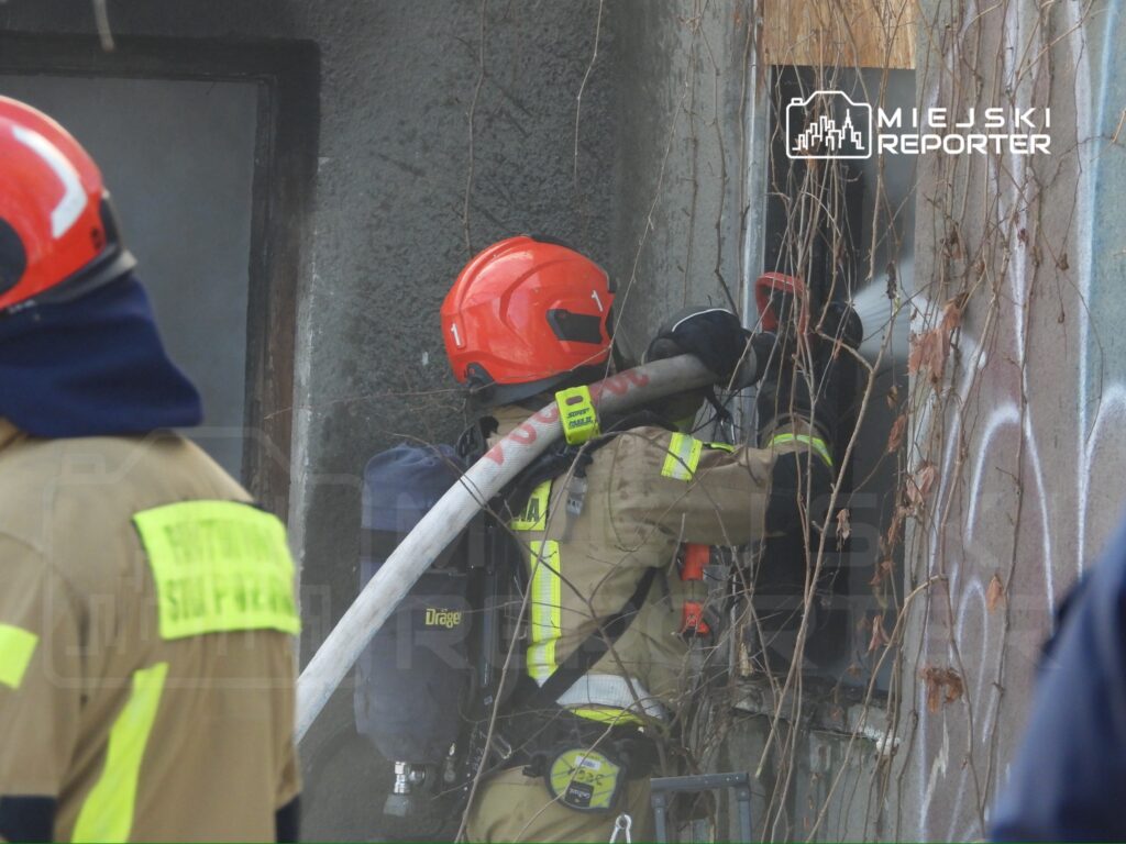 Strażak w hełmie i mundurze używa węża gaśniczego, gasząc ogień w oknie budynku z graffiti.