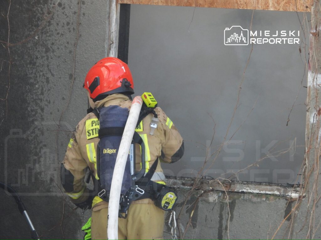 Strażak w hełmie i mundurze gaszącym pożar przy oknie budynku, trzymający wąż gaśniczy.