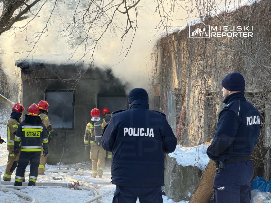 Grupa strażaków w hełmach i mundurach gasi pożar w budynku, podczas gdy policjanci monitorują sytuację.