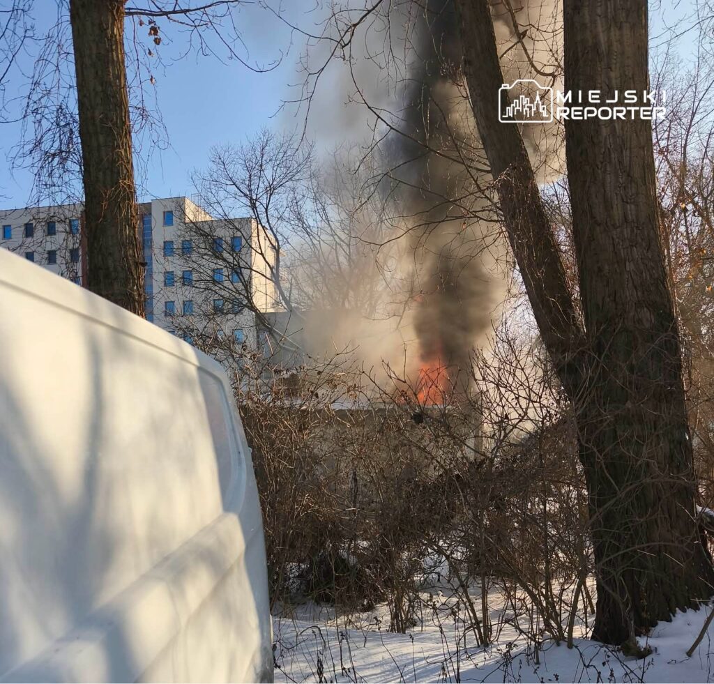W tle nowoczesny budynek, z którego wydobywa się czarny dym, a w pobliżu widoczny biały samochód.