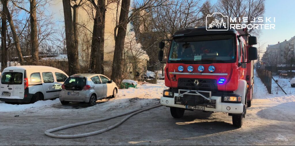 Czerwony wóz strażacki z włączonymi sygnalizatorami stoi na zaśnieżonym terenie, obok zaparkowanych samochodów.