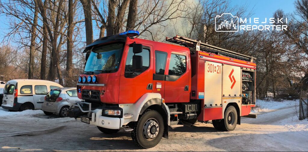 Czerwony wóz strażacki zaparkowany na śnieżnej drodze, w tle drzewa i inne pojazdy.