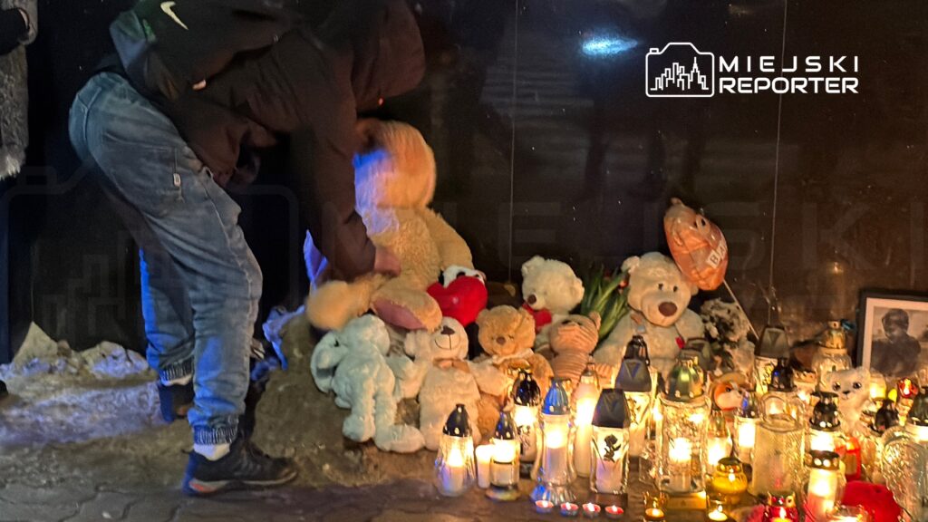 Mężczyzna w czarnej kurtce układa pluszowe misie obok świec w hołdzie zmarłemu.