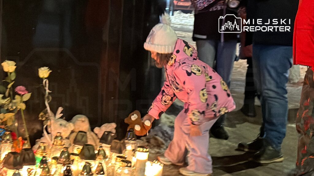 Dziewczynka w różowej bluzie z postacią z bajki kładzie pluszowego misia obok zniczy i kwiatów w ciemnym otoczeniu.