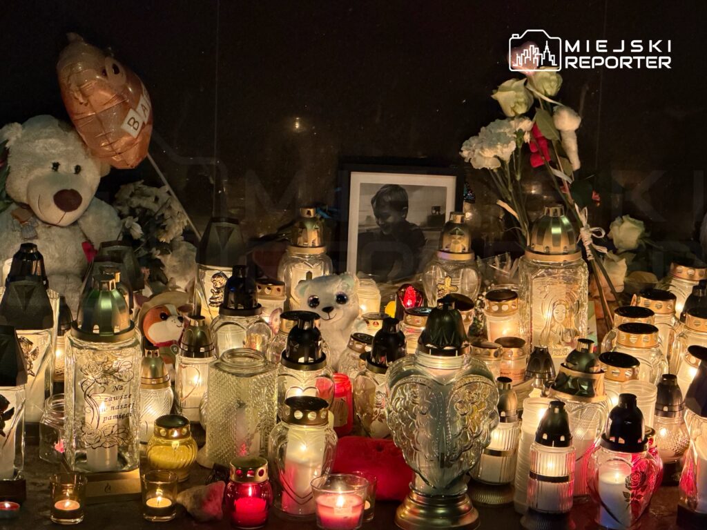 Na oświetlonym miejscu znajdują się znicze, pluszowe misie i zdjęcie dziecka, otoczone kwiatami.