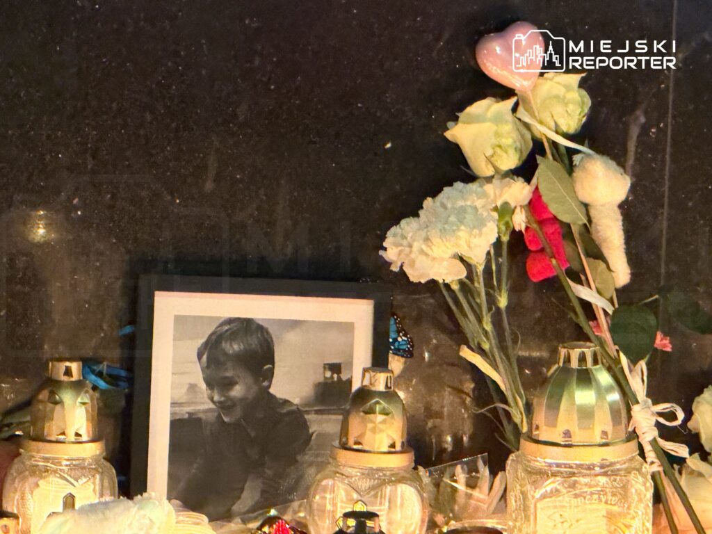 Cztery znicze oraz kwiaty stoją obok zdjęcia uśmiechającego się chłopca na ciemnym tle.
