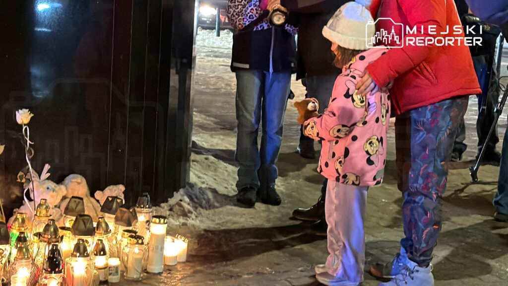 Dzieci z lampionami stoją przed zniczami ustawionymi na chodniku, w tle widać ciemny budynek.