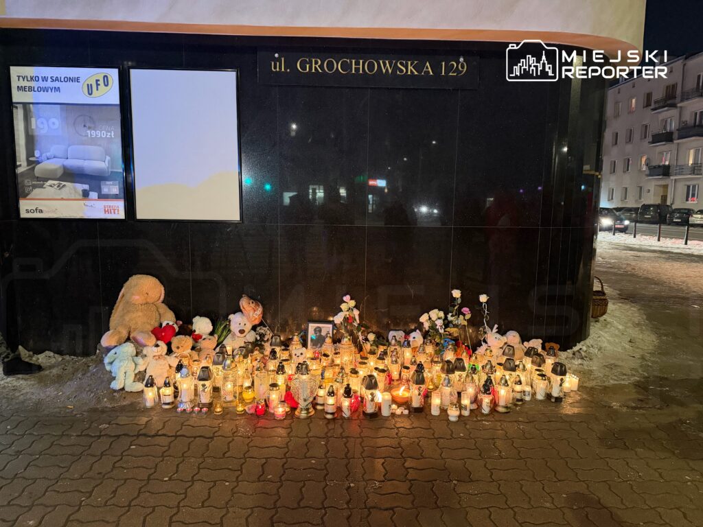 Zbiór zniczy i pluszaków ustawionych obok ciemnej ściany budynku w miejskim otoczeniu.