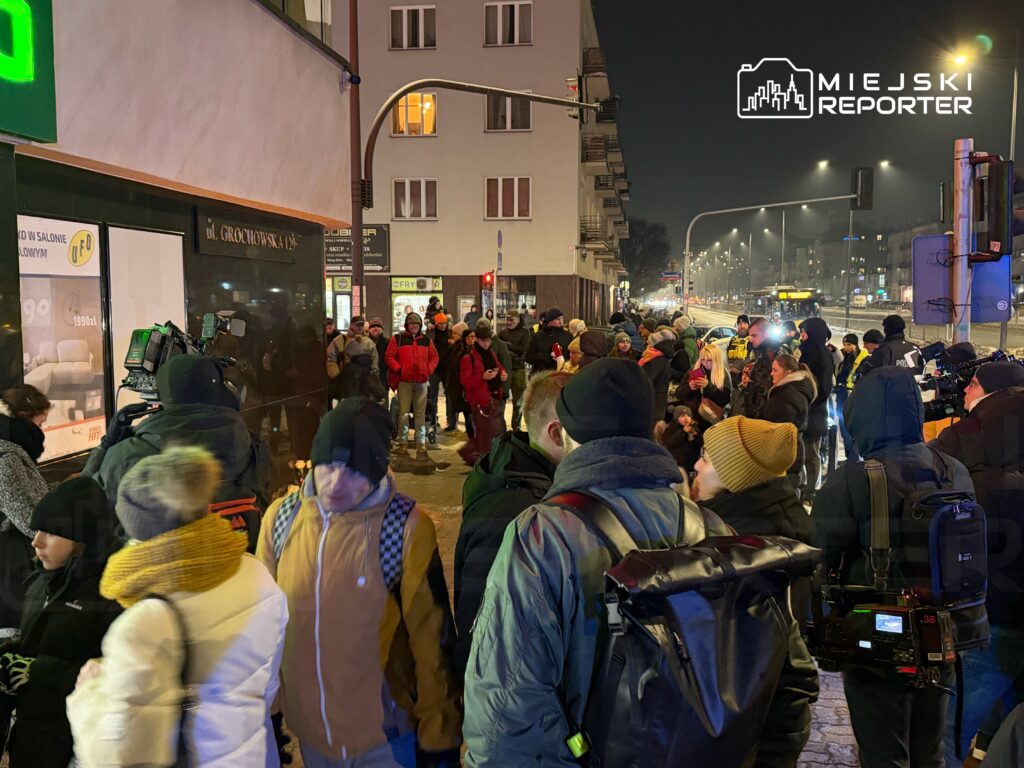 Tłum ludzi w zimowych ubraniach gromadzi się na chodniku, a w tle widoczne są kamery telewizyjne i oświetlenie uliczne.