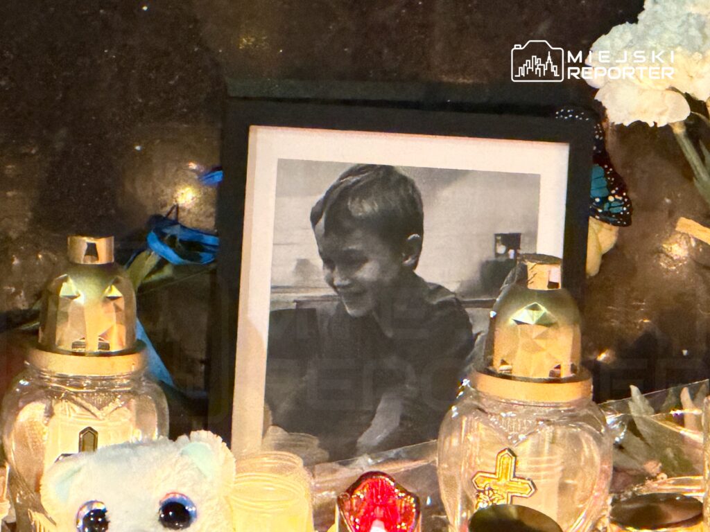 Czarna ramka z czarno-białym zdjęciem uśmiechniętego chłopca otoczona zniczami i pluszowym misiem.