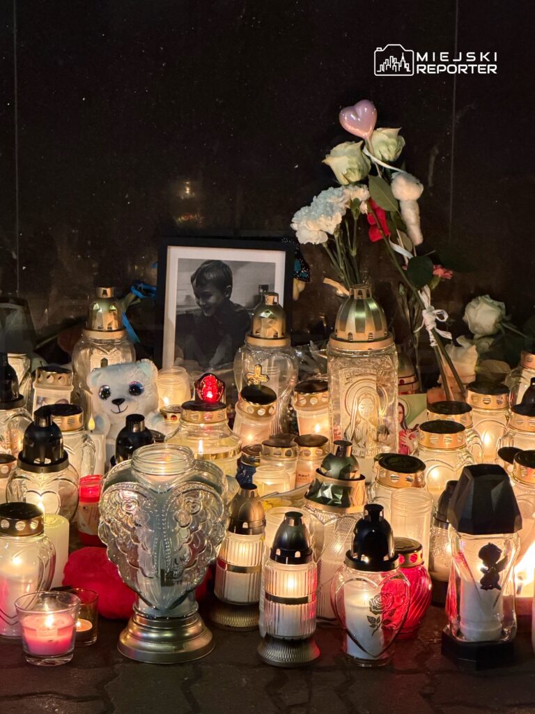 Zgromadzenie zniczy i kwiatów przy zdjęciu dziecka, w otoczeniu świec i pamiątek, na ciemnym tle.