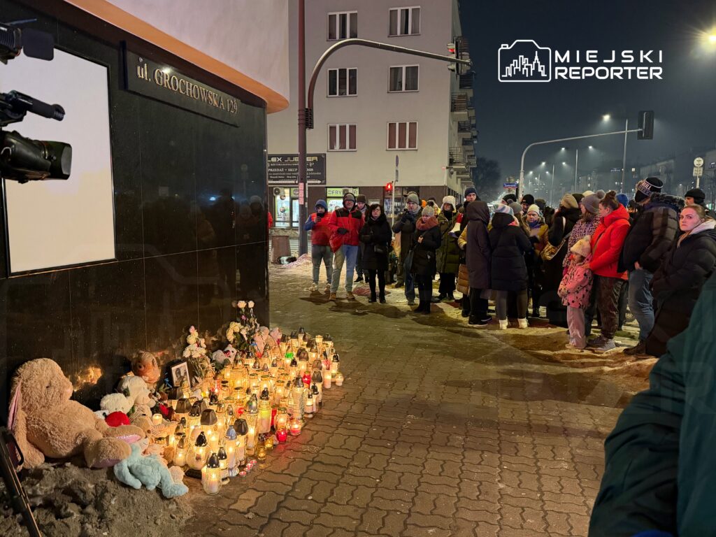 Grupa ludzi stoi w kolejce na chodniku, obok zniczy i pluszowych zabawek ustawionych przed ciemnym murem budynku.