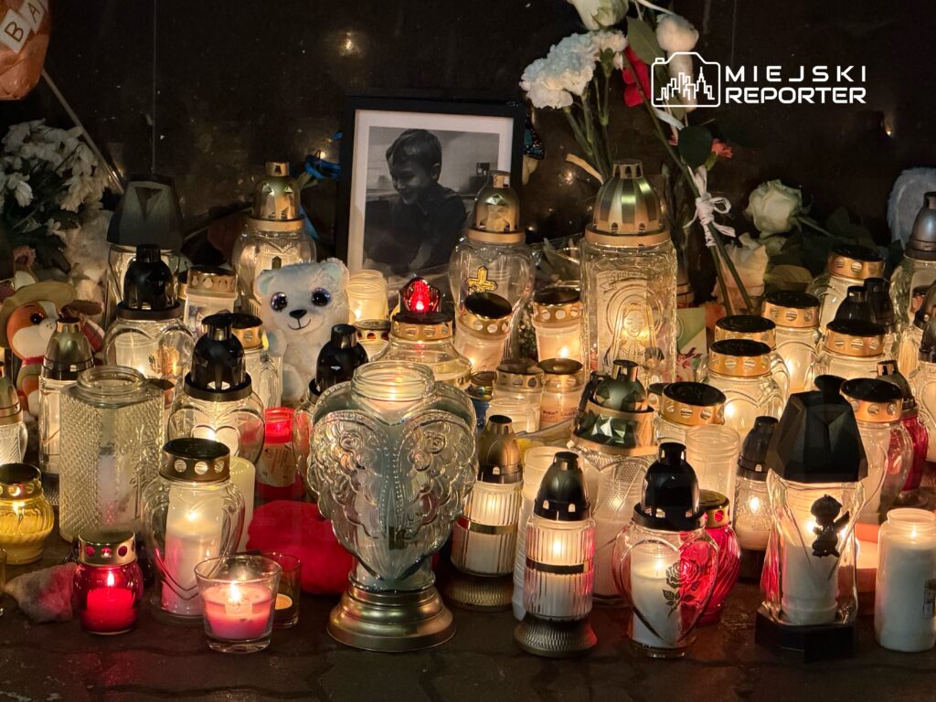 Świeczki w różnych kształtach i kolorach otaczają zdjęcie uśmiechniętego chłopca, obok leżą kwiaty.