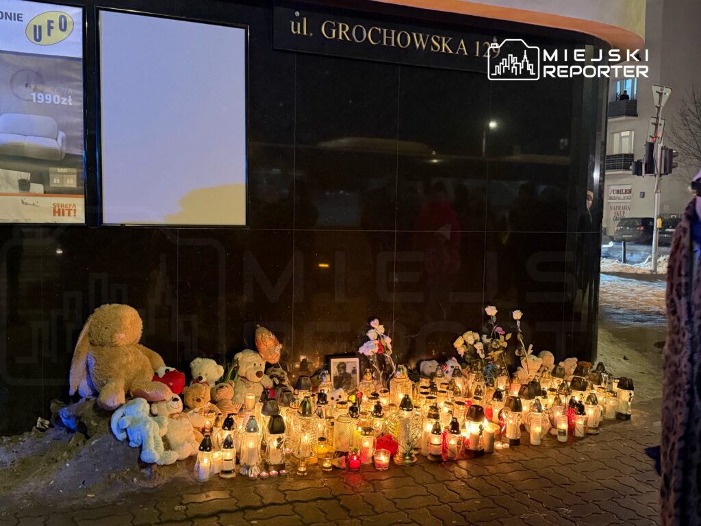 Miejsce pamięci z pluszowymi zabawkami i zniczami ustawionymi przy czarnej ścianie budynku.