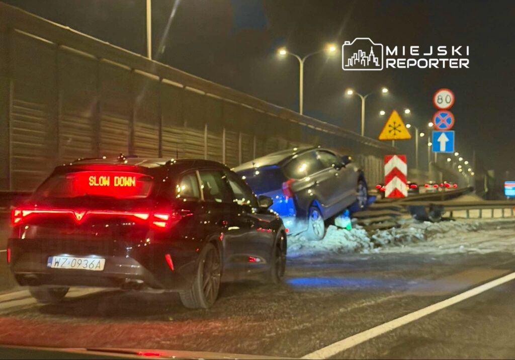 Czarny samochód z wyświetlaczem "SLOW DOWN" zatrzymany obok drogi, obok unieruchomiony pojazd na poboczu.